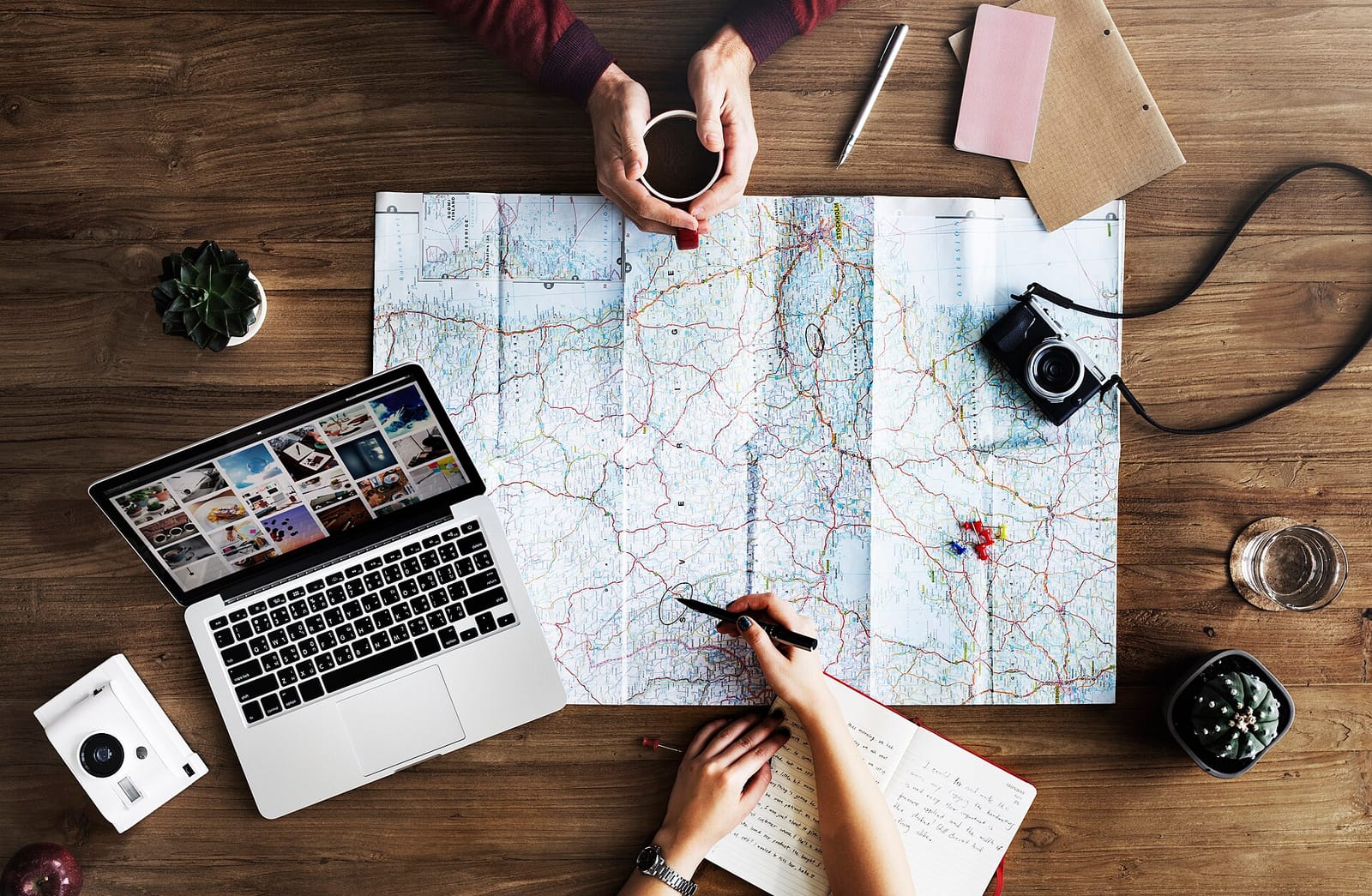 People planning a trip around a paper map, with a laptop, camera and coffee on a wooden table